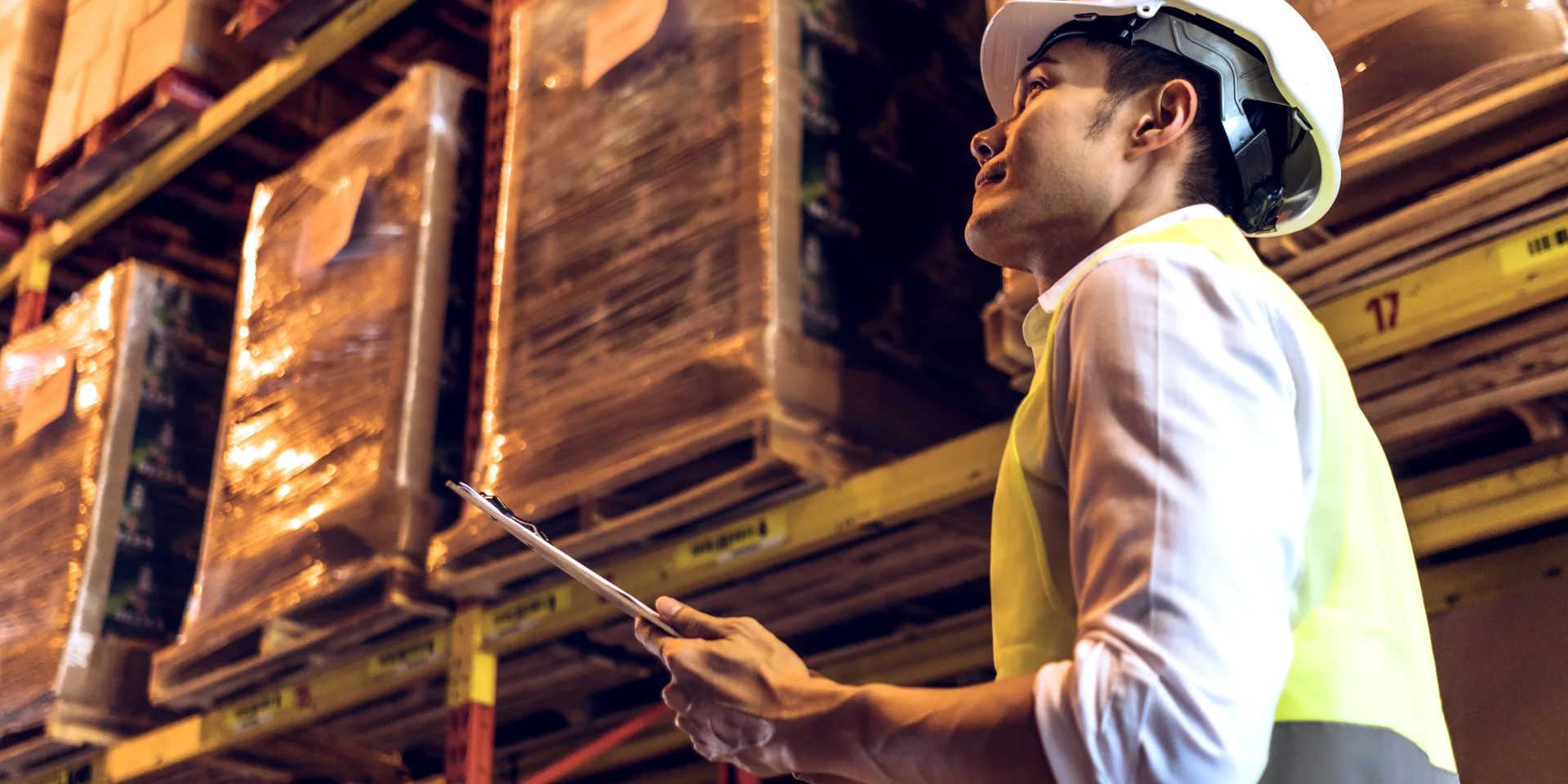 A warehouse worker checking stock against a clipboard beneath racked pallets